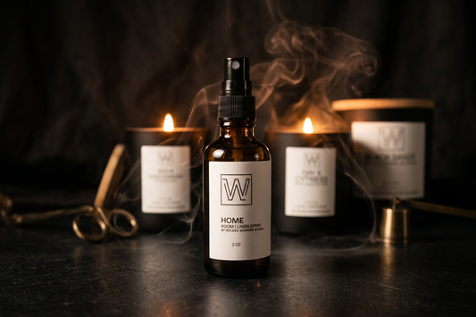 An amber glass home fragrance spray bottle with a black spray top and white label stands in the center, surrounded by three lit candles and a diffuser jar in the background with visible smoke. The labels show a large stylized W logo and the word HOME near the bottom.