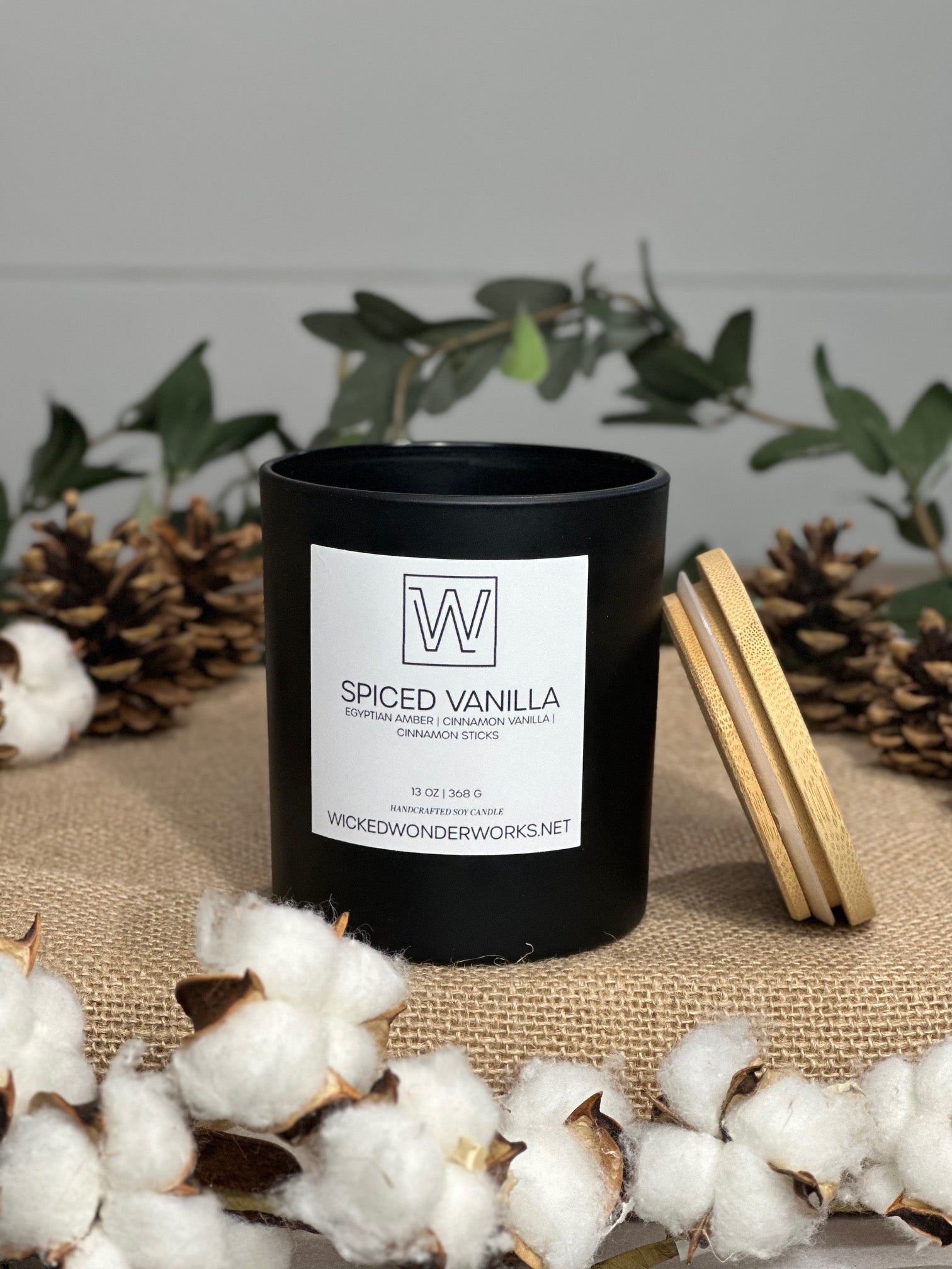A black soy candle with a label reading 'Spiced Vanilla' placed on a burlap surface with pine cones and decorative leaves around it.