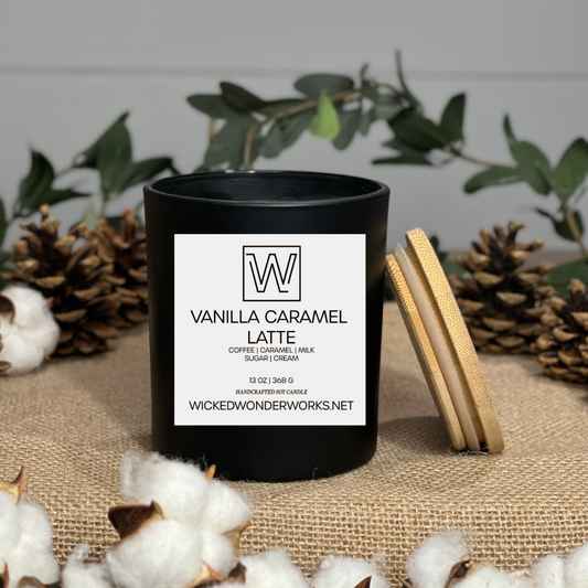 Black candle with a vanilla caramel latte label on a textured surface with greenery and cotton flowers.
